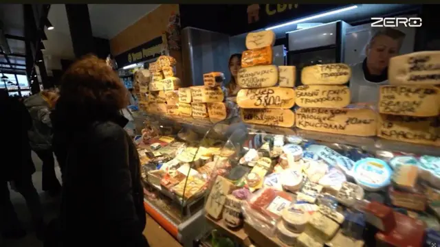 На рынке перед стеклянной витриной с сырными головками стоит покупательница в темной куртке. На полках – десятки сортов сыра с рукописными ценниками; за прилавком видны холодильные шкафы. Внутри зала много света и людей.