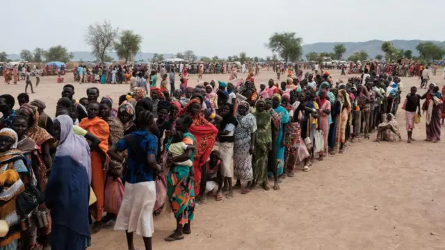 Des femmes et des enfants forment une longue file d'attente, tandis que de nombreux autres se tiennent en groupes dans une zone ouverte et poussiéreuse. Seuls quelques abris temporaires épars sont visibles.