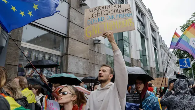 Pessoas na marcha do Orgulho, incluindo um homem segurando uma placa que diz "Soldados LGBTQ ucranianos defendem nossa liberdade - dê-lhes seus direitos"