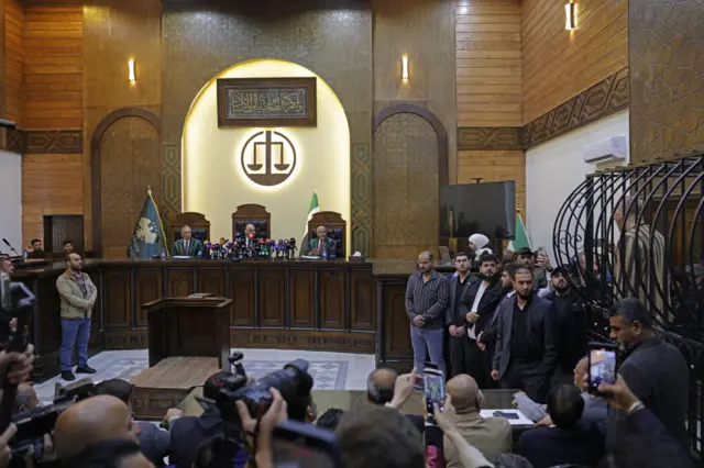 أشحاص يقفون داخل قاعة محكمة وفي الخلفية قفص الاتهام، بين يجلس ثلاثة قضاة، وأمامهم حاضرون.