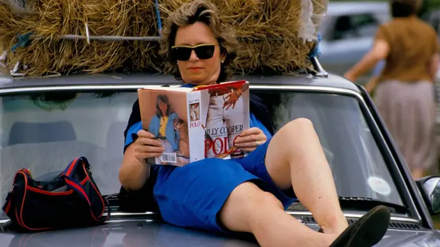 Жінка читає книгу Джиллі Купер, сидячи зверху на капоті авто