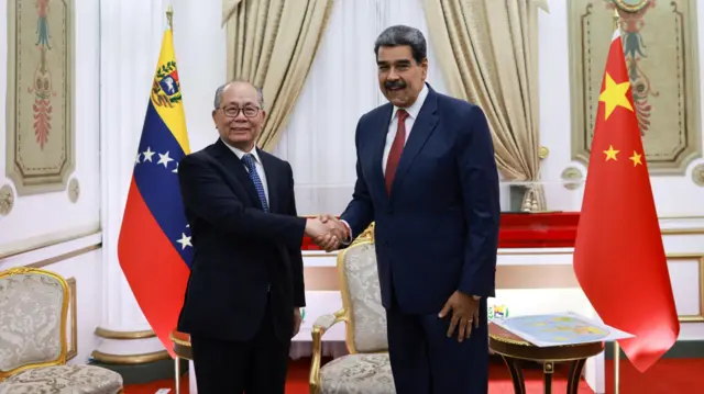 Nicolás Maduro cumprimenta Qiu Xiaoqi no Palácio Miraflores, sede do governo venezuelano em Caracas. Ambos estão sorrindo, usam ternos escuros e gravata, com as bandeiras da Venezuela e da China ao fundo.