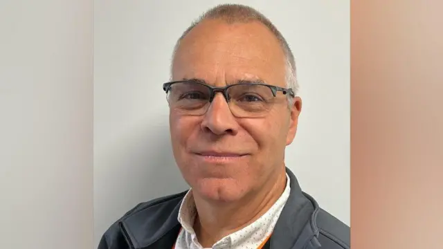 Richard has very short dark grey hair and wears glasses. He is wearing a white shirt with small blue dots on it, and a grey jacket
