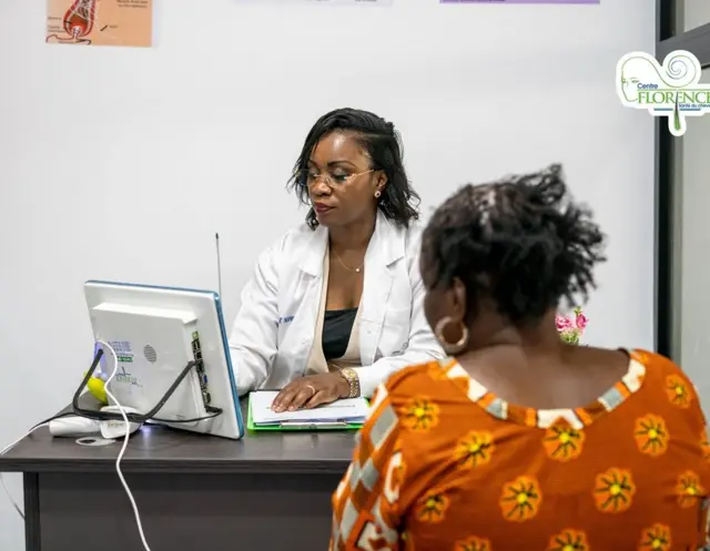 Dr Florence Edwige Nanga en blouse blanche assise dans son bureau en traint de regarder l'écran de son ordinateur, elle est en consultation avec une patiente en robe à motif
