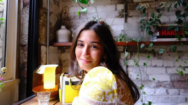 Mulher de cabelos castanhos e camiseta amarela sorri dentro de uma cafeteria com luz baixa.