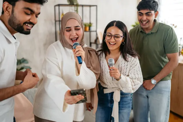 দুই জন পুরুষ ও দুই জন নারী মাইক্রোফোন হাতে একটি মোবাইল ফোন দেখে গান গাইছেন