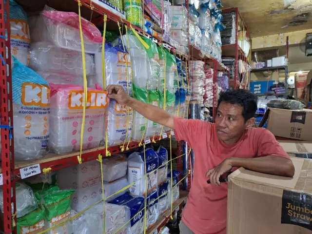 Ronny dengan kaos berkerah merah sedang menunjukkan tumpukan wadah plastik di warungnya.