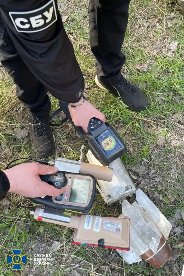 На траві лежать уламки невеликої ракети. У кадрі видно двох людей у чорному одязі з нашивками «СБУ», які тримають портативні прилади для вимірювання — один із них має дисплей із показником близько 58,5 (ймовірно, рівень випромінювання). Прилади прикладені безпосередньо до уламків.