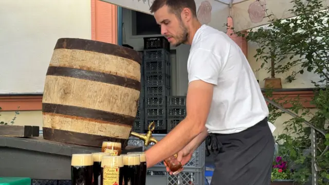 Barman nalewa ciemne piwo z beczki. Ma na sobie białą koszulkę i fartuch, a jego zdjęcie zrobione jest z profilu. Na stoliku obok niego stoi około sześciu piw.