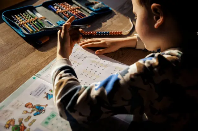 Menino escrevendo em caderno infantil; ele segura caneta com a mão esquerda 