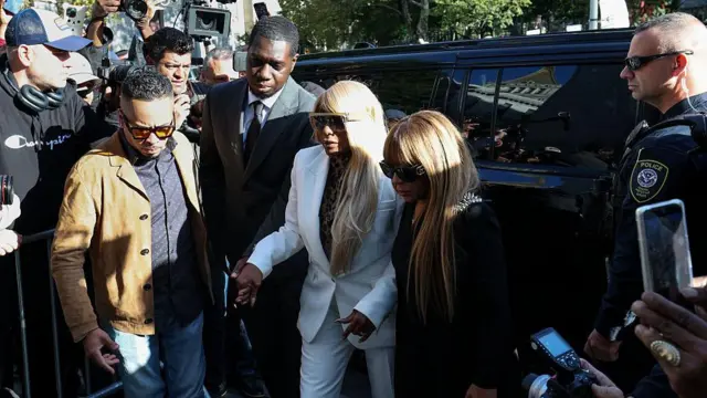 Джанис Комбс в белом брючном костюме и темных очках выходит из машины в сопровождении своего антуража.