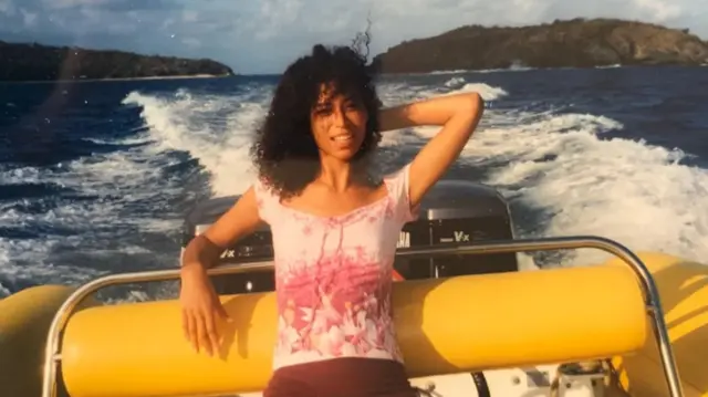 Lisa Philips on a boat wearing a pink top and with her left arm posing behind her head. Epstein's island is in the background