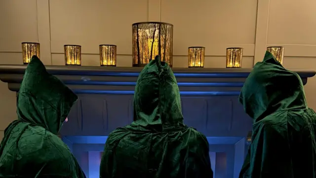 Las personas visten capuchas de terciopelo verde y miran hacia una chimenea gris. Sobre la chimenea hay siete faroles dorados con siluetas de árboles.