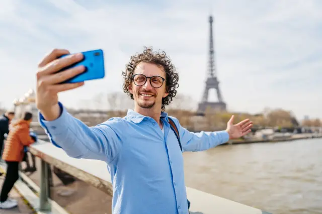 Homem tirando selfies com seu smartphone em uma ponte em Paris, na França. Vista da Torre Eiffel ao fundo.
