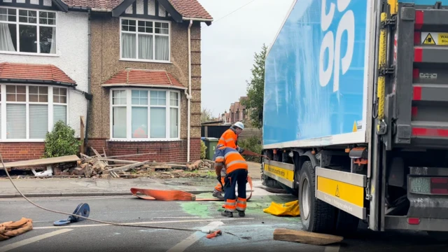 Dos trabajadores vestidos con uniformes de color naranja brillante limpian los escombros de una carretera junto a un camión azul después de un choque con un automóvil, con madera dañada apilada afuera de una casa adosada que anteriormente era parte de una cerca.