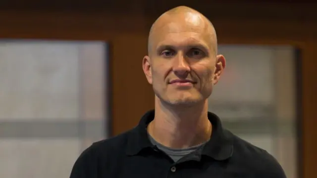 Profeseri Jason Stearns ahagaze areba muri 'camera', yambaye umupira muremure w'umukara. Imbere yambayemo undi mupira ujya kuba icyatsi kibisi. 