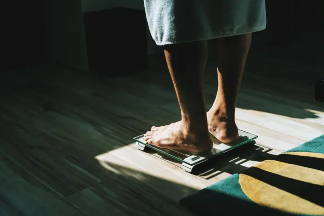 Людина стоїть босоніж на підлогових вагах у кімнаті з дерев’яною підлогою; на ногах — рушник або халат, світло падає з боку, створюючи тіні.
