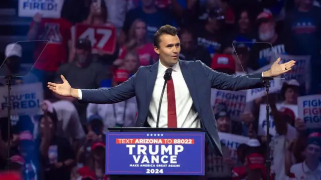 Uma imagem de Charlie Kirk discursando em um comício de Trump em Glendale, Arizona, em agosto de 2024. Atrás dele, uma multidão vestida com roupas vermelhas, brancas e azuis, segurando cartazes com os dizeres "VOCÊ ESTÁ DEMITIDO" e "LUTE! LUTE! LUTE!". Ele veste um terno azul com uma camisa branca de botões e uma gravata vermelha, discursando em um pódio enquanto gesticula amplamente com as mãos. No pódio, há uma placa com os dizeres "TRUMP-VANCE, FAÇAM A AMÉRICA GRANDE NOVAMENTE EM 2024".