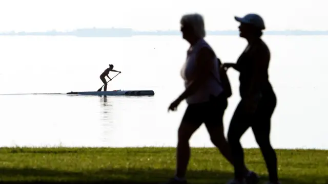 Dwie kobiety spacerują, w tle na wodzie inna kobieta płynie na desce z wiosłem