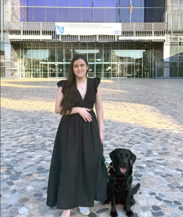 Una mujer con vestido negro, de pie junto a un perro lazarillo negro.