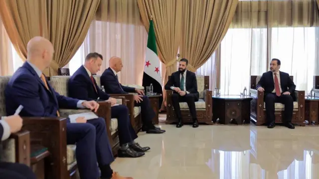 Barrack ve heyeti Şara ve dışişleri bakanıyla birlikte koltuklarda oturuyor, arada Suriye bayrağı var.