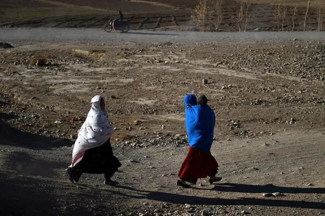 دو زن افغان در حال راه رفتن در یک جاده خاکی. یکی از آنها کودکی در آغوش دارد.