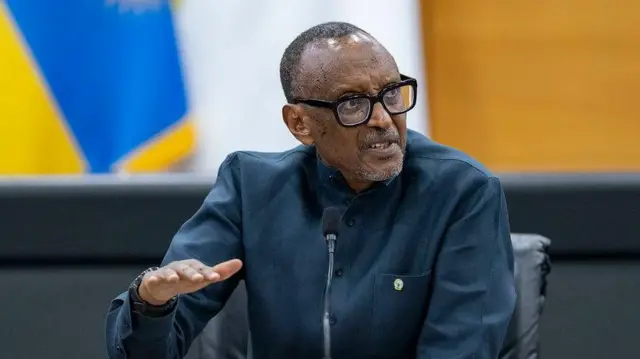Perezida w'u Rwanda Paul Kagame mu kiganiro n'abanyamakuru ku wa kane, ku itariki ya 27 Ugushyingo (11) mu 2025, mu biro bye Village Urugwiro, i Kigali, mu Rwanda. Yicaye mu ntebe y'umusego w'umukara, yambaye indorerwamo n'ishati yijimye y'amaboko maremare, akoze ikimenyetso n'ikiganza. Imbere ye hari indangururamajwi arimo kuvugiramo, iriho agapamba k'umukara. Arareba ku ruhande. 