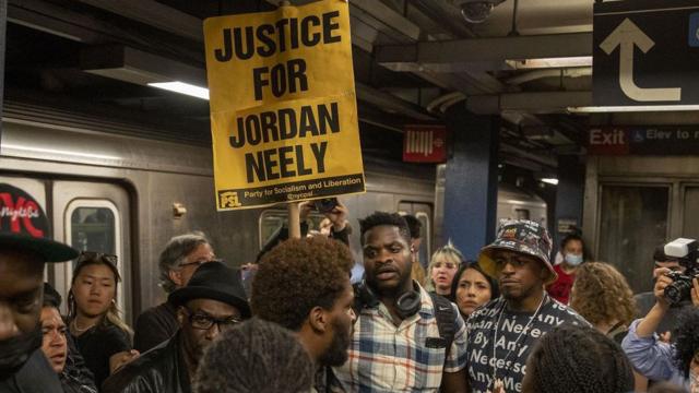 Plataforma do metrô cheia, com manifestante erguendo cartaz que diz: Justiça para Jordan Neely