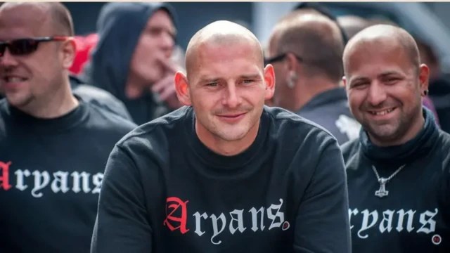 سه نفر با لباس یک شکل سیاه رنگ که روی آن نوشته شده آریایی‌ها و سرهای طاس به دوربین نگاه می‌کنند و لبخند می‌زنند پشت سر آنها هم چند نفر با لباس یک شکل 