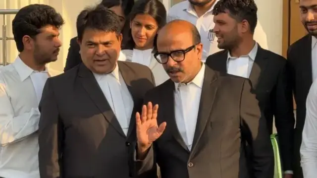 ਕਾਲੇ ਕੋਟ ਪਹਿਨੀ ਦੋ ਵਕੀਲ ਅਤੇ ਪਿੱਛੇ ਕੁਝ ਹੋਰ ਵਕੀਲ ਨਜ਼ਰ ਆ ਰਹੇ ਹਨ