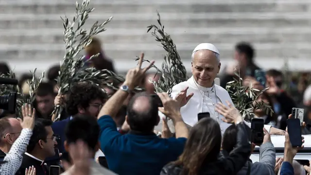 Понтифик в белом облачении на папамобиле среди толпы людей, у некоторых в руках мобильные телефоны или пальмовые ветви.