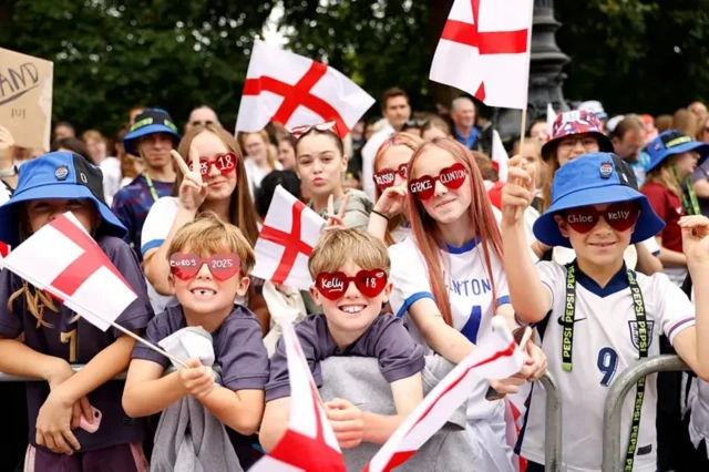 Люди с флагами и с очками в виде сердечек, на которых написаны имена футболисток. Многие из собравшихся дети