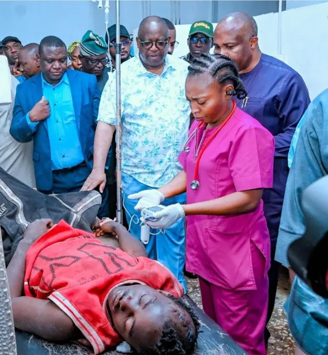 Gomina Caleb Mutfwang ti ipinlẹ Plateau wọ asọ shaati ati sokoto alawọ buluu, n fi ọwọ kan ọkunrin kan to wọ asọ pupa,di irun sori, sun sori ibusun nile iwosan. Obinrin Nọọsi kan to wọ ibọwọn duro niwaju gomina.