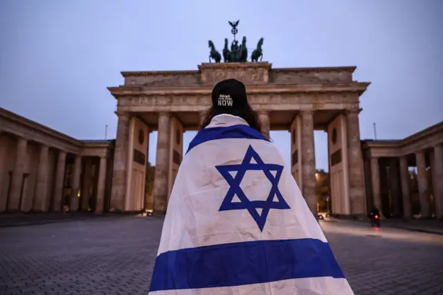 Человек, завернутый в израильский флаг, сфотографирован на фоне Бранденбургских ворот в Берлине в сумерках
