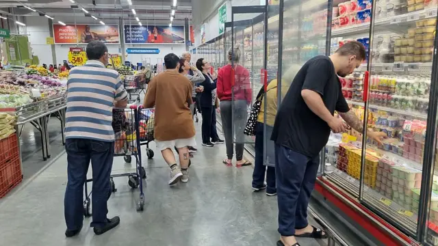 Pessoas no corredor de um supermercado, com gôndolas de vegetais à esquerda e geladeiras de laticínios à esquerda