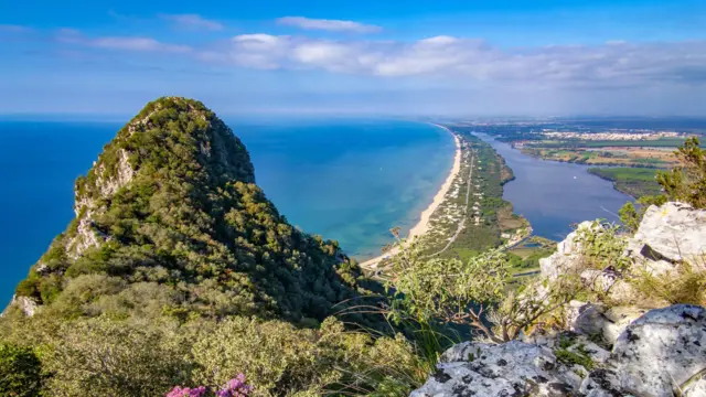 Widok z Monte Circeo na wybrzeże Riwiery Odyseusza we Włoszech. Na pierwszym planie widać porośnięty zielenią skalisty szczyt góry, a w tle długą piaszczystą plażę oraz laguny i równiny ciągnące się wzdłuż wybrzeża.