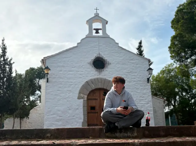 Alex Batty está sentado con las piernas cruzadas sobre un muro bajo de piedra, sosteniendo un teléfono móvil, con una botella de plástico para bebidas colocada a su lado.