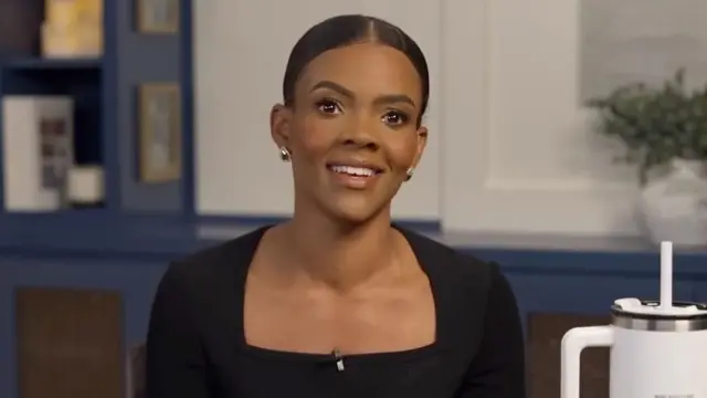 Candace Owens usa blusa preta de gola quadrada e brincos pequenos e dourados, enquanto sorri para frente. O fundo da imagem é uma parede azul escura, com quadros pendurados e uma planta em vaso à direita. 