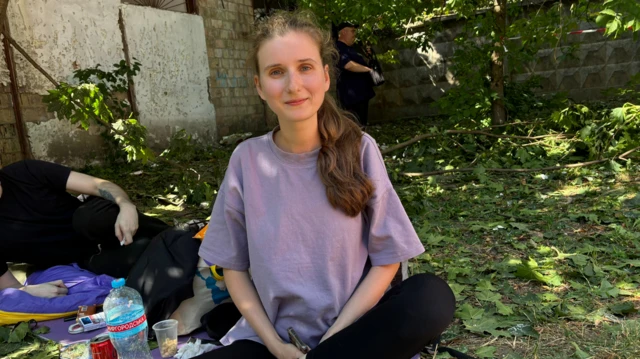 Анастасия Ковтун ерда оёқларини чалиштириб ўтирибди, атрофида барглар ётибди, орқа фонда эса бир эркак ён бош ётган ҳолда кўринмоқда.