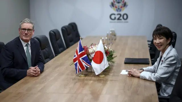 施紀賢爵士（左）與高市早苗（右）在南非約翰內斯堡G20峰會週邊會晤，兩人坐在長會議桌中央兩側，桌面中間擺放著英國與日本小型國旗（22/11/2025）