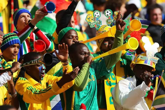 South Africa football fans wearing team colours, a headband, woolly and hard hats, blow multicoloured vuvuzelas at a match.
