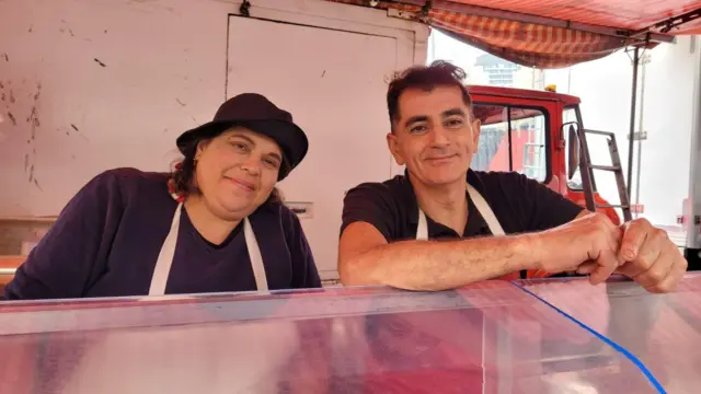 A foto mostra uma mulher e um homem, ambos de camiseta escura e um avental branco, atrás de um balcão.