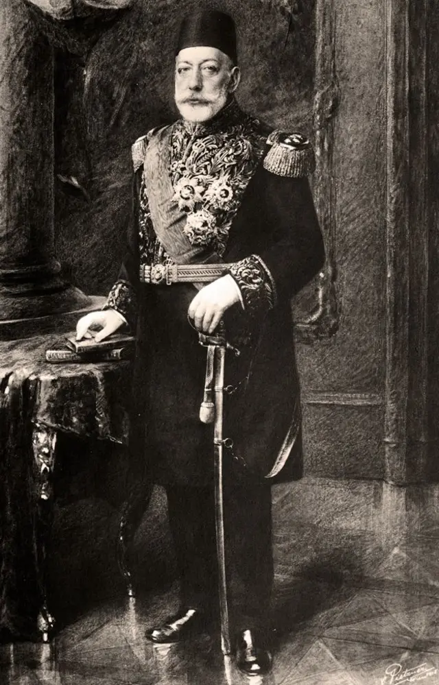 Mehmed V Reshad, de barba blanca, está parado al lado de una mesa. Una de sus manos reposa sobre unos libros y la otra sostiene lo que parece ser una espada que toca el suelo. Viste un elegante traje militar y lleva puesta un tarboosh