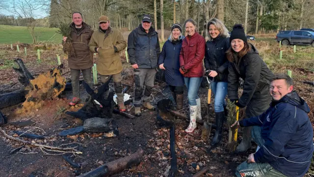 Un pequeño grupo de personas sonrientes, todas con abrigos de invierno, sonríe a la cámara junto a un trozo de tierra excavada. Dos de ellas sostienen palas de jardinería, y otra sostiene lo que parece ser un pequeño arbusto o árbol perenne. Al fondo se ve una zona de bosque y dos camiones.