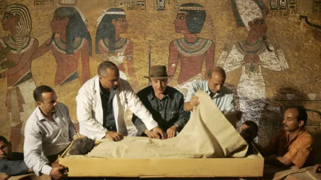 Con la ayuda de técnicos en mangas de camisa y bata de laboratorio, el jefe de antigüedades de Egipto, Zahi Hawass (centro), con un sombrero fedora marrón al estilo de Indiana Jones, supervisa la extracción de la momia de Tutankamón, envuelta en lino, de su sarcófago de piedra, en su tumba subterránea, en el Valle de los Reyes, en Luxor, el 4 de noviembre de 2007. Arte egipcio antiguo, que muestra figuras con cabezas y piernas de perfil pero torsos de frente, y jeroglíficos adornan la pared detrás.