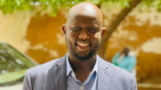 Mohamed Dore, souriant face à la caméra, vêtu d'une chemise bleue à carreaux et d'un blazer bleu marine.