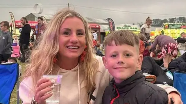 Нікола і Зак на фестивалі. Вони усміхаються в камеру. Нікола тримає пластиковий стаканчик, обіймаючи Зака