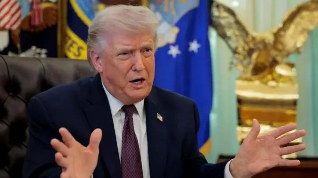 さまざまな旗や金色の彫刻を前に、机に向かいながら両手を広げて話すトランプ氏