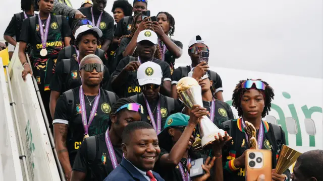 Les joueuses camerounaises, vêtues pour la plupart de t-shirts noirs et arborant des médailles d'or suspendues à des rubans violets autour du cou, se tiennent sur les marches d'un avion tandis que le trophée de la Coupe d'Afrique des nations féminine est brandi bien haut. On aperçoit, devant les joueuses au pied de l'escalier, le visage souriant d'un homme en costume.