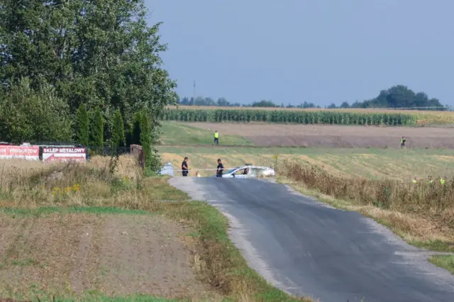 Поліцейська автівка посеред поля, огороджена стрічкою. У полі, на тлі густої кукурудзи, видно людей у зелених жилетках